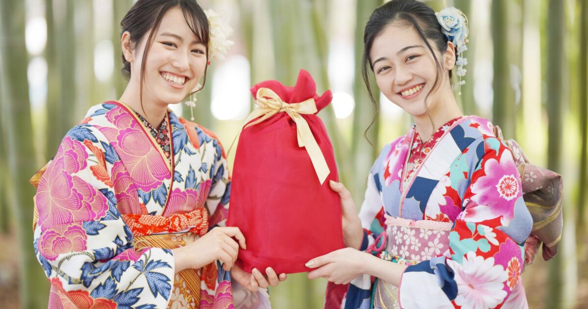着物姿の女性二人が赤い風呂敷包みを持って笑顔を見せる様子